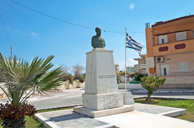 La bandiera greca sventola a fianco a un monumento commemorativo, con busto. 